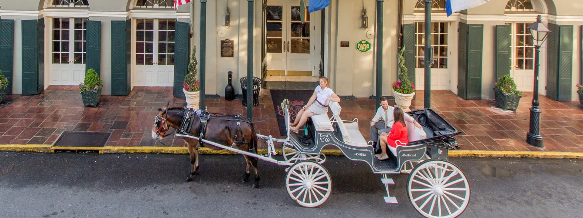 A horse-drawn carriage with people is in front of a building with a balcony and columns.