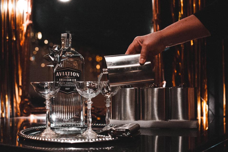 A hand pouring a drink from a metal shaker into a glass, with a bottle of Aviation American Gin and two glasses on a tray.