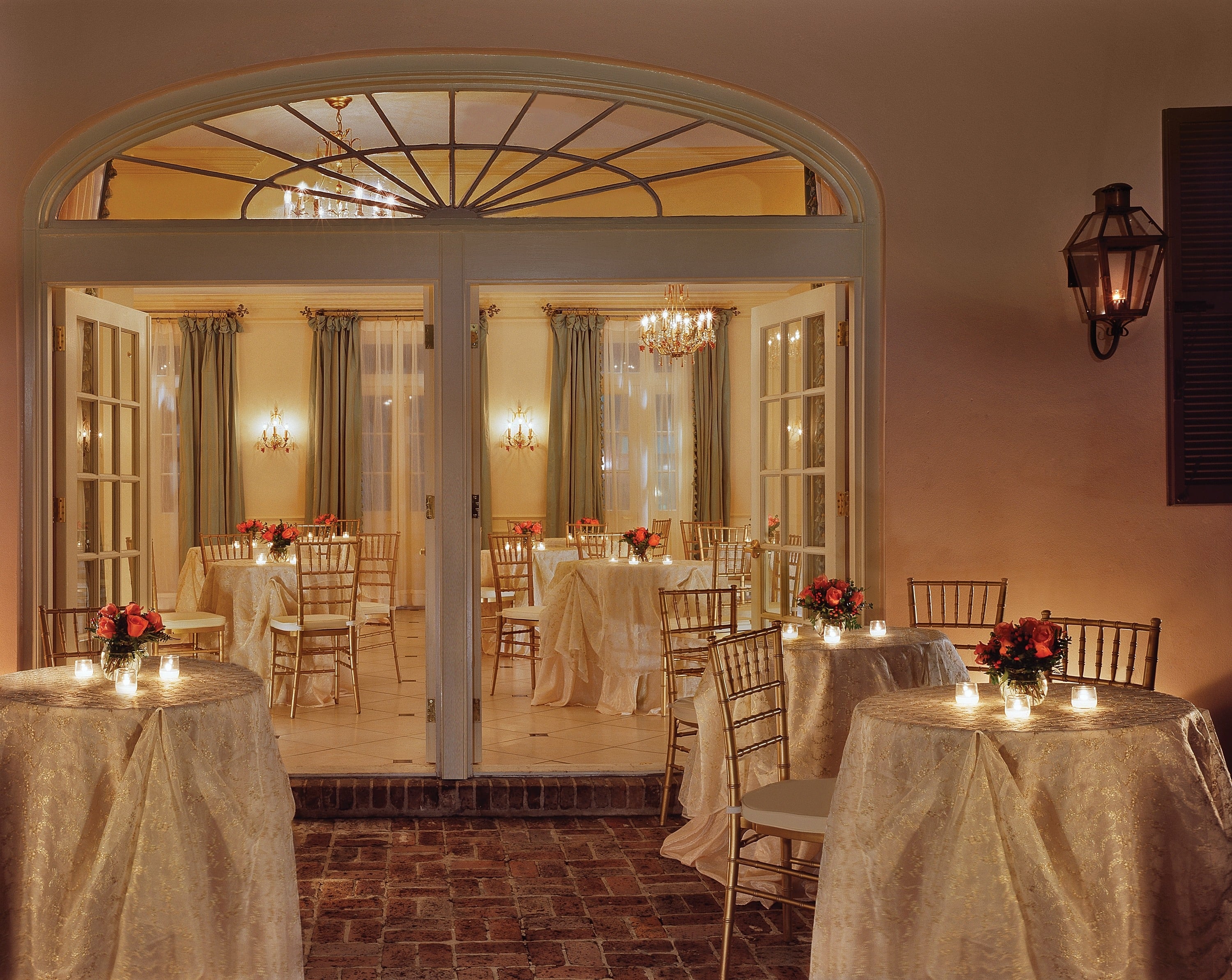 A formal dining room with round tables, white tablecloths, red flowers, and candles, with glass doors leading to another room.
