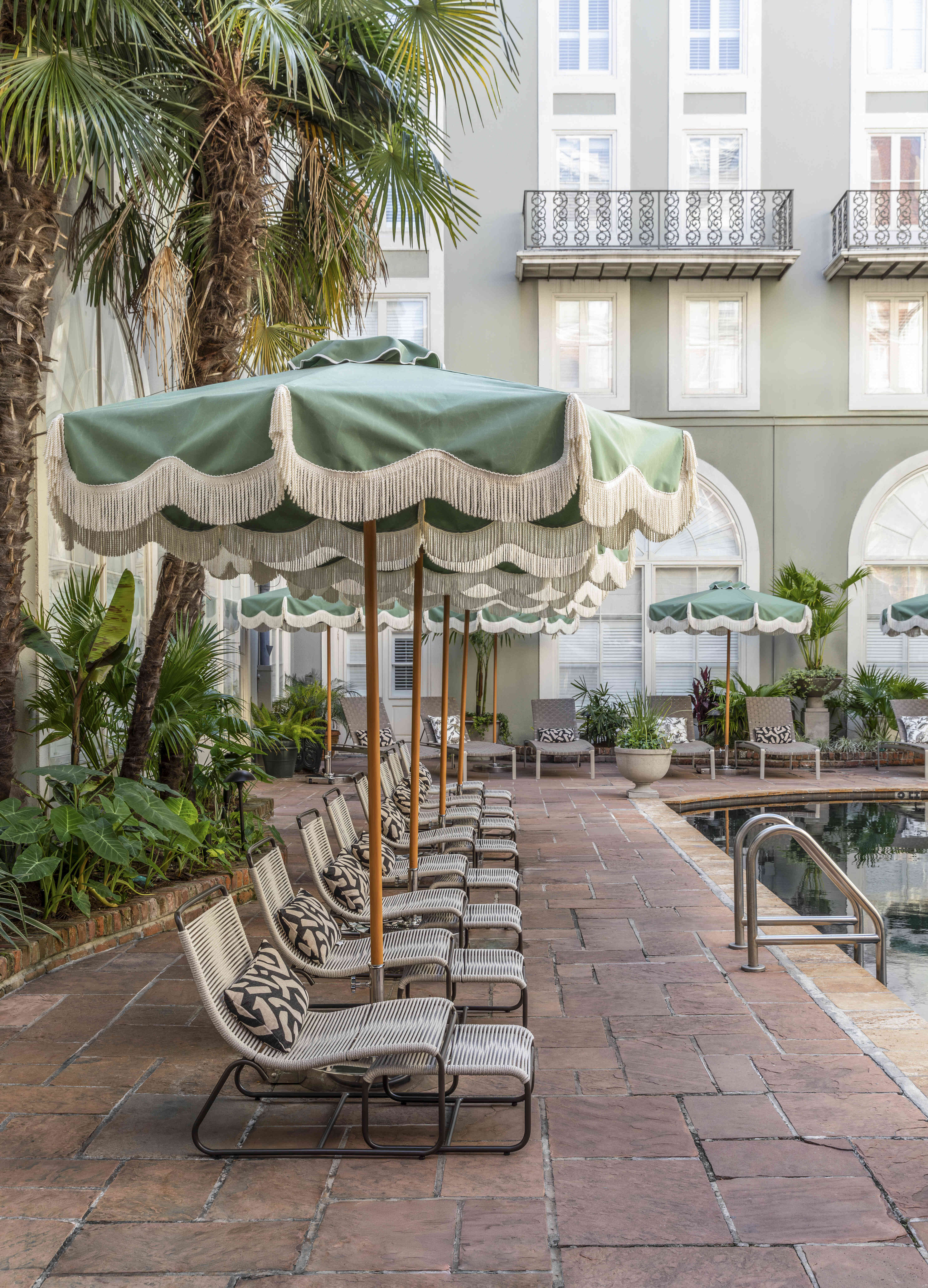 A poolside area with green umbrellas, lounge chairs, and palm trees in front of a building with balconies.
