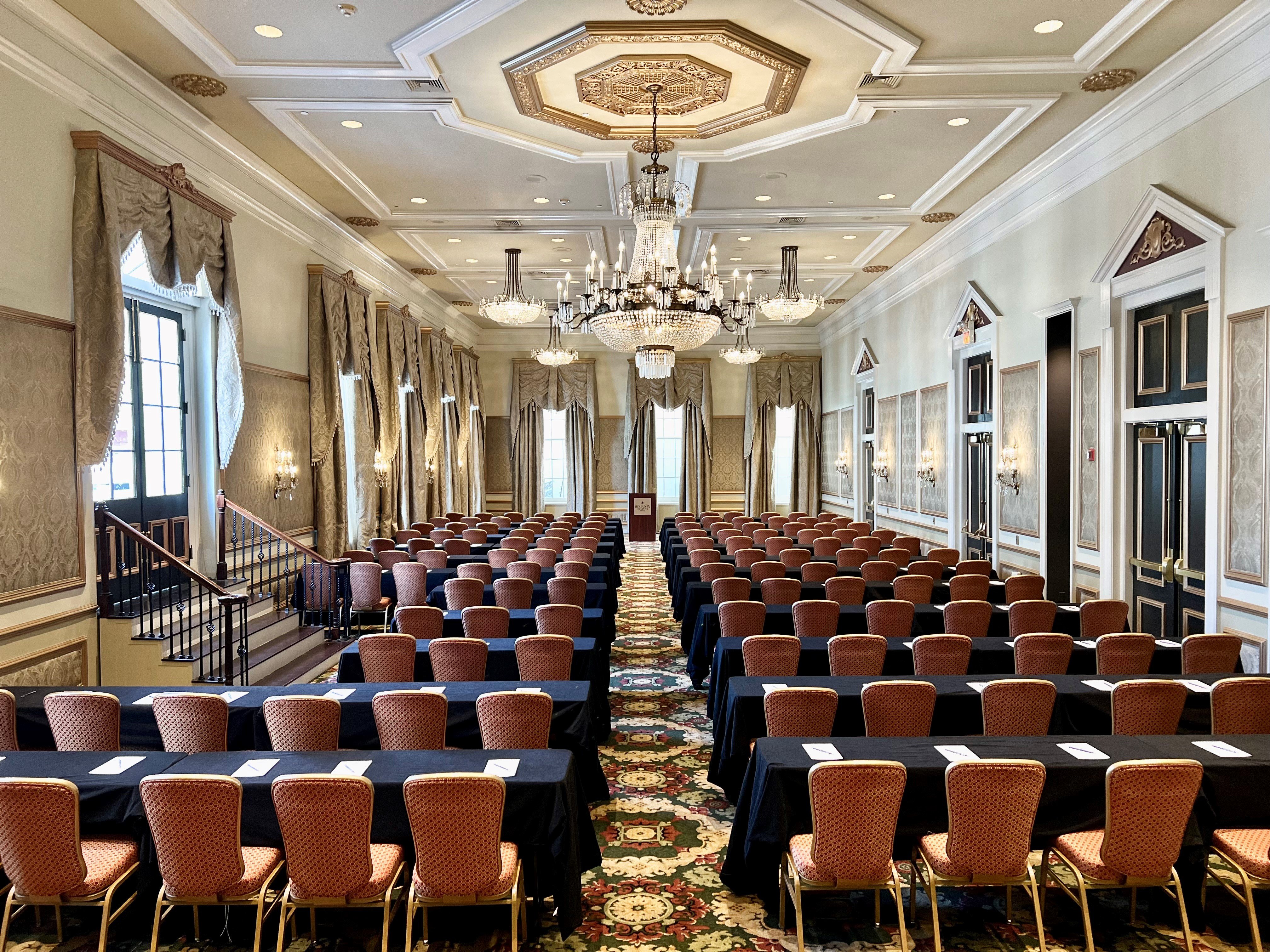 A large, ornate meeting room with rows of tables and chairs, a chandelier, and curtains.