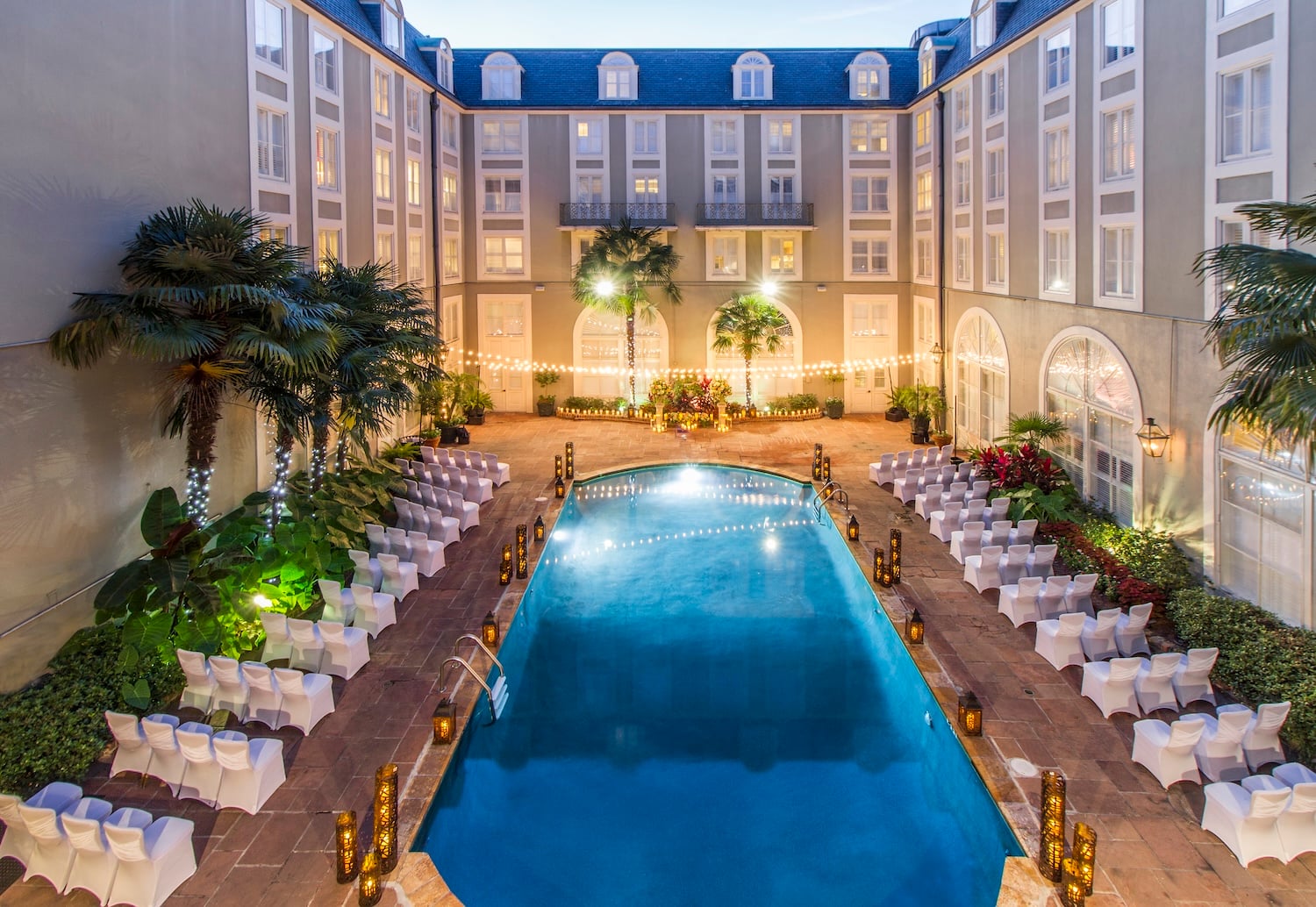 A pool surrounded by white chairs and palm trees in a courtyard with buildings in the background.
