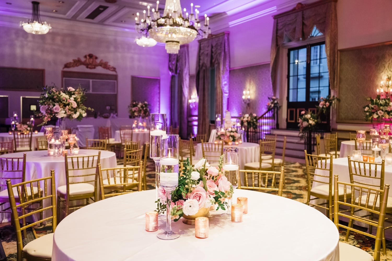 A lavishly decorated banquet hall with round tables, white tablecloths, gold chairs, floral arrangements, and candles.