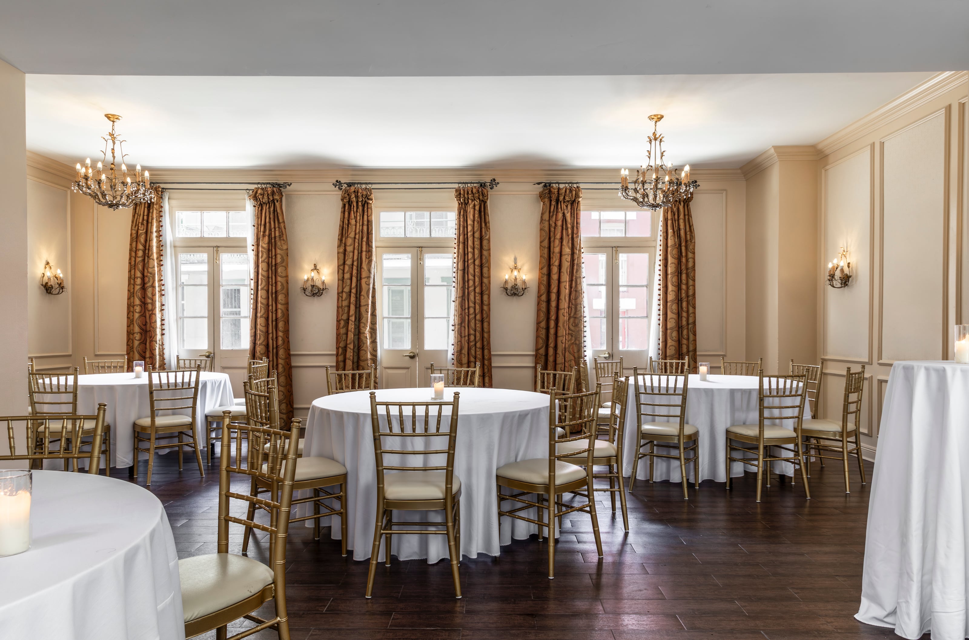 A formal dining room with round tables, white tablecloths, gold chairs, chandeliers, and large windows.